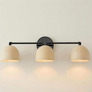Peut inclure: Une applique murale à trois lumières composée d'une barre horizontale en métal noir et d'une fixation murale circulaire noire. Trois abat-jours en forme de dôme beige mat sont fixés à la barre, chacun abritant une ampoule diffusant une lumière chaude.