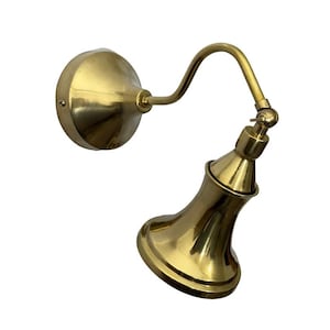 May include: A polished brass wall sconce featuring a circular mounting plate, a curved arm, and a bell-shaped shade. The fixture includes an adjustable joint at the top of the shade, allowing for directional light positioning with a metallic finish.