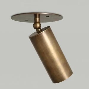 May include: A bronze-coloured, cylindrical spotlight fixture. The light is attached to a circular base and can be angled. The fixture is designed for ceiling mounting and has a modern, minimalist aesthetic.