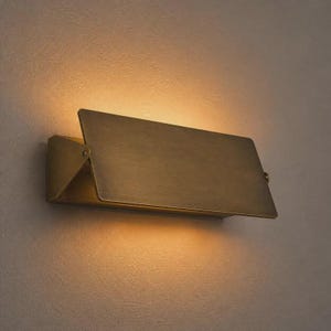 Pode incluir: Um candeeiro de parede retangular com acabamento em latão escovado. A luminária possui um painel frontal plano e inclinado que projeta luz tanto para cima quanto para baixo, criando um brilho quente e suave na superfície da parede.