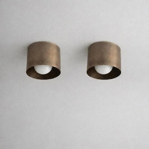 May include: Two cylindrical flush mount light fixtures featuring a brushed bronze metal exterior and a central white glass globe bulb. The design is simple and circular, with the metallic housing surrounding the light source on all sides.