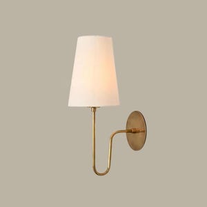Op de afbeelding: Een wandlamp met een witte lampenkap en een messing afwerking. De lamp heeft een gebogen arm en een cirkelvormige achterplaat. De lampenkap is taps toelopend en geeft een warme gloed. De armatuur is ontworpen voor binnenverlichting.