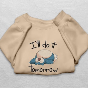 May include: A beige crewneck sweatshirt featuring an embroidered graphic of a blue and cream-colored creature lying down with its eyes closed. The text "I'll do it tomorrow" is embroidered in black lettering above and below the character.