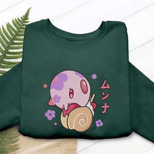 Peut inclure: Un sweat-shirt &agrave; col rond vert fonc&eacute; orn&eacute; d'un motif repr&eacute;sentant une cr&eacute;ature rose et ronde &agrave; fleurs violettes pos&eacute;e sur un g&acirc;teau roul&eacute; beige. Le dessin inclut des petites fleurs violettes, une fraise et le texte japonais ムンナ en rouge.