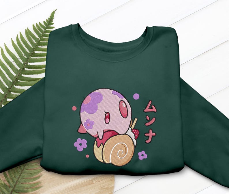 Peut inclure: Un sweat-shirt &agrave; col rond vert fonc&eacute; orn&eacute; d'un motif repr&eacute;sentant une cr&eacute;ature rose et ronde &agrave; fleurs violettes pos&eacute;e sur un g&acirc;teau roul&eacute; beige. Le dessin inclut des petites fleurs violettes, une fraise et le texte japonais ムンナ en rouge.