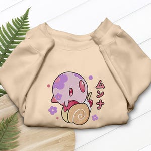 Peut inclure: Un sweat-shirt &agrave; col rond beige orn&eacute; d'un motif repr&eacute;sentant une cr&eacute;ature ronde violet clair &agrave; motifs floraux, pos&eacute;e sur un g&acirc;teau roul&eacute; beige surmont&eacute; d'une fraise. Le design inclut de petites fleurs violettes et le texte japonais ムンナ.