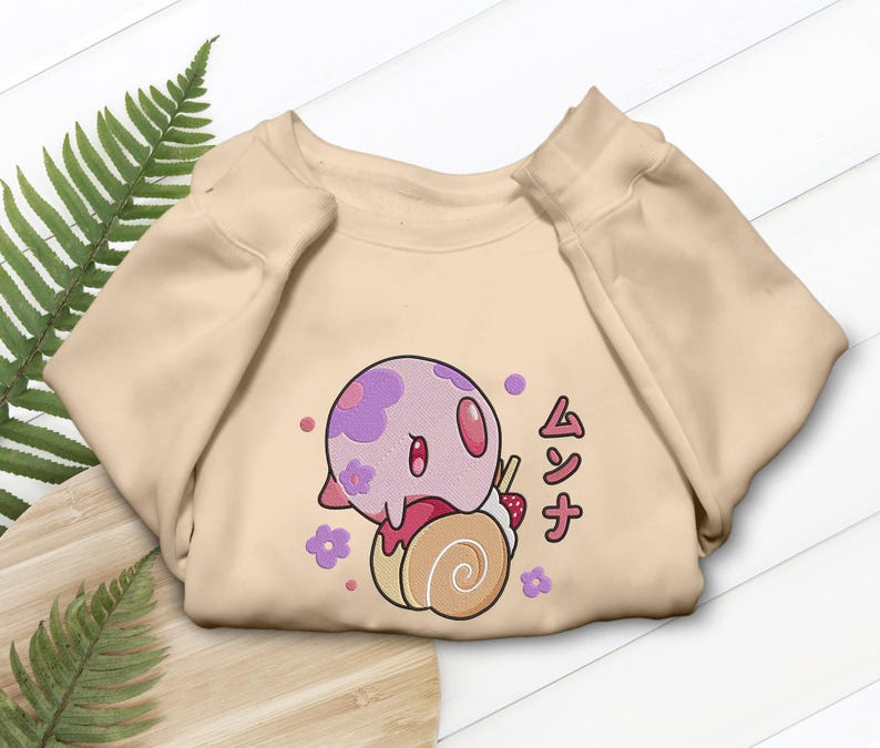 Peut inclure: Un sweat-shirt &agrave; col rond beige orn&eacute; d'un motif repr&eacute;sentant une cr&eacute;ature ronde violet clair &agrave; motifs floraux, pos&eacute;e sur un g&acirc;teau roul&eacute; beige surmont&eacute; d'une fraise. Le design inclut de petites fleurs violettes et le texte japonais ムンナ.