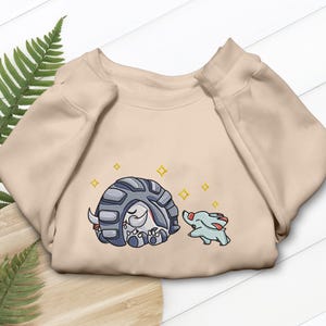 May include: A beige crewneck sweatshirt featuring a graphic print of a large, grey, armored creature curled into a ball next to a smaller, light blue creature with red accents. Yellow four-pointed star shapes are scattered around the figures.