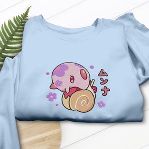 Peut inclure: Un sweat-shirt bleu clair avec une broderie repr&eacute;sentant une cr&eacute;ature rose et ronde &agrave; motifs floraux violets pos&eacute;e sur une p&acirc;tisserie en spirale beige. Le motif inclut des petites fleurs violettes, des points roses et le texte japonais ムンナ en rouge.
