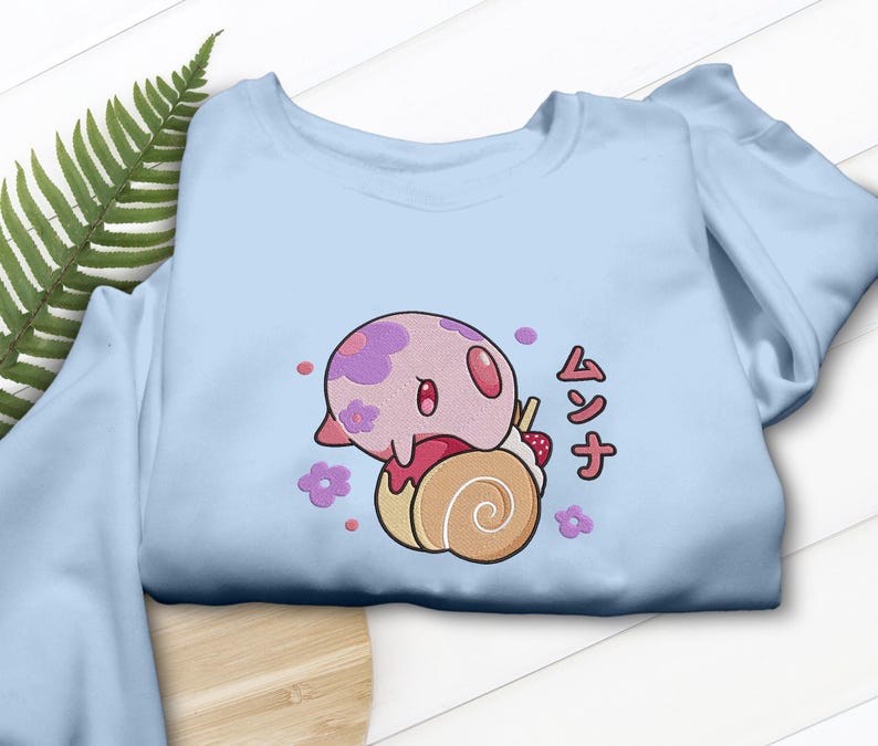 Peut inclure: Un sweat-shirt bleu clair avec une broderie repr&eacute;sentant une cr&eacute;ature rose et ronde &agrave; motifs floraux violets pos&eacute;e sur une p&acirc;tisserie en spirale beige. Le motif inclut des petites fleurs violettes, des points roses et le texte japonais ムンナ en rouge.