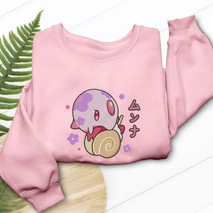 Peut inclure: Un sweat-shirt &agrave; manches longues rose clair orn&eacute; d'un motif repr&eacute;sentant une cr&eacute;ature ronde &agrave; taches violettes pos&eacute;e sur une p&acirc;tisserie beige en forme de spirale avec une fraise. Le dessin inclut des fleurs violettes et le texte japonais ムンナ.