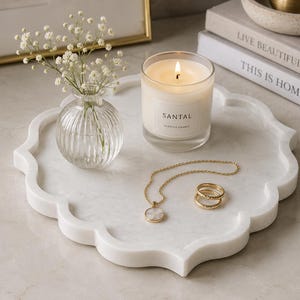 Könnte beinhalten: Ein weißes Marmortablett mit gewelltem Rand, darauf eine gerippte, durchsichtige Glasvase mit weißen Schleierkrautblüten, eine brennende Kerze im Glas mit der Aufschrift Santal, eine goldene Halskette mit Anhänger und zwei goldene Ringe.
