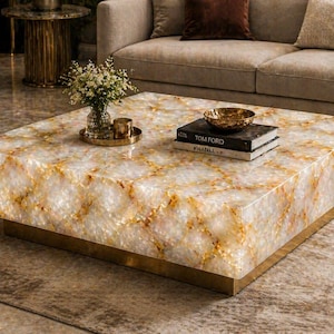 May include: A square coffee table with a white and gold patterned top and a gold base. The table is accessorized with a small vase of flowers, a decorative bowl, and two books.