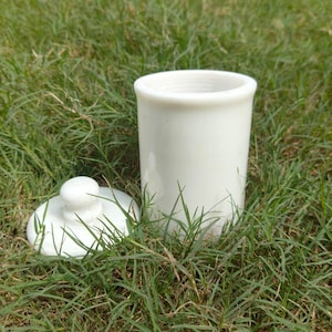 May include: A cylindrical white ceramic canister with a matching circular lid featuring a small knob handle. The container has a smooth, solid white finish and a simple, functional design, presented as two separate pieces resting side by side.