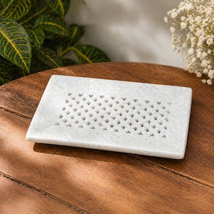 May include: A rectangular, white marble soap dish with a grid of small, heart-shaped cutouts. The dish is set on a dark wooden surface, with green foliage and white flowers in the background. The soap dish is approximately 6 inches long and 4 inches wide.