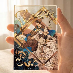 Puede incluir: Bloque rectangular de acrílico transparente con una ilustración detallada de tres figuras con túnicas y sombreros, sobre un fondo de elementos celestiales y detalles dorados. La obra tiene una temática fantástica.