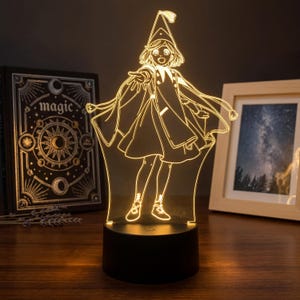 Puede incluir: Lámpara LED 3D con diseño de bruja de dibujos animados. La lámpara tiene una base negra y una figura de acrílico transparente de una bruja con sombrero y capa. Un libro con la palabra "magic" es visible en el fondo.