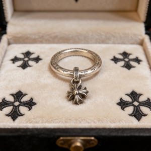 May include: A silver ring with a dangling cross pendant sits in a black jewellery box with a cream velvet lining. The box is decorated with four black cross designs. The ring is detailed with small embellishments.