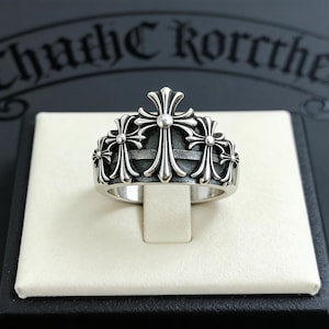 May include: A silver ring with a cross design. The ring features multiple crosses arranged around the band. The ring is displayed on a white surface, with a dark background. The ring has a detailed, ornate design.