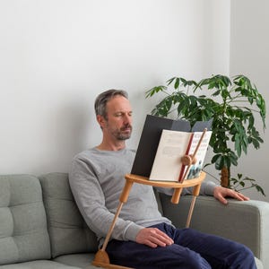 Könnte beinhalten: Ein hölzerner Buchständer mit natürlichem Finish, der ein offenes Buch hält. Der Ständer hat ein kleines Regal und verstellbare Beine. Die Person sitzt auf einem grauen Sofa und liest. Eine grüne Pflanze ist im Hintergrund zu sehen.