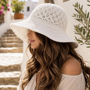 May include: A white crochet sun hat with a wide, slightly downturned brim and a textured, intricate floral-inspired pattern across the crown. The hat is constructed from a woven material, featuring a consistent off-white colour throughout the design.