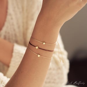 May include: Three delicate cord bracelets featuring small gold-toned charms. The top bracelet has a star charm, the middle one has a cross charm on a dark cord, and the bottom one has a heart charm. The bracelets have adjustable cord ends.