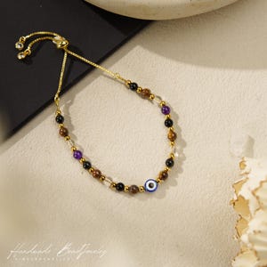 May include: A gold-tone adjustable bracelet featuring a series of multi-colored beads including black, brown, purple, and clear stones, accented with small gold-tone metal spacers and a single blue and white evil eye bead charm.