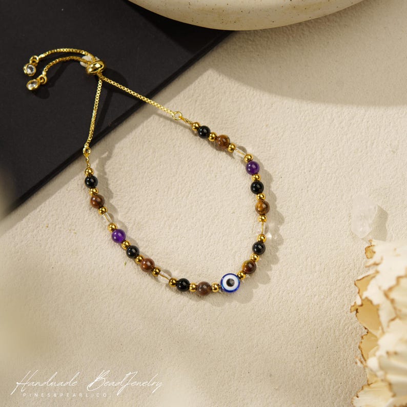 May include: A gold-tone adjustable bracelet featuring a series of multi-colored beads including black, brown, purple, and clear stones, accented with small gold-tone metal spacers and a single blue and white evil eye bead charm.