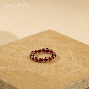 May include: A ring constructed from a continuous series of small, deep red spherical beads. Each individual bead is secured in place by thin, metallic gold-coloured wire that wraps around the circumference to form a circular band structure.