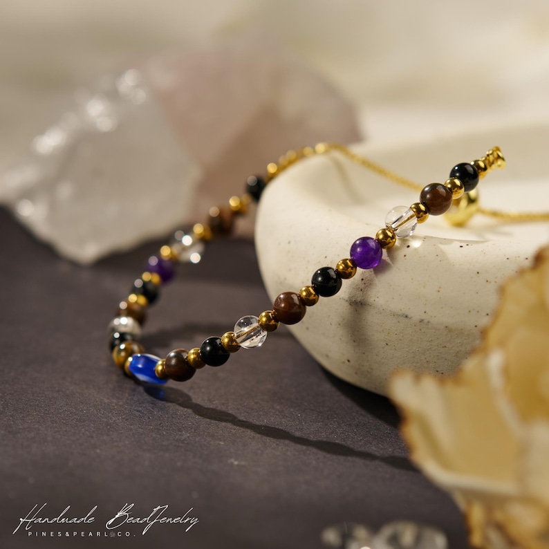May include: A beaded bracelet featuring a sequence of small, polished round stones in shades of black, brown, purple, blue, and clear, interspersed with tiny metallic gold-toned beads. The bracelet includes a thin gold-toned chain component.