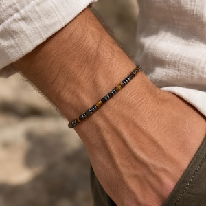 Puede incluir: Una pulsera fina de cuentas con un patrón repetitivo de pequeñas cuentas cilíndricas de color gris metálico oscuro y piedras de ojo de tigre de color marrón dorado. La pulsera presenta un diseño sencillo con un cierre plano y seguro.