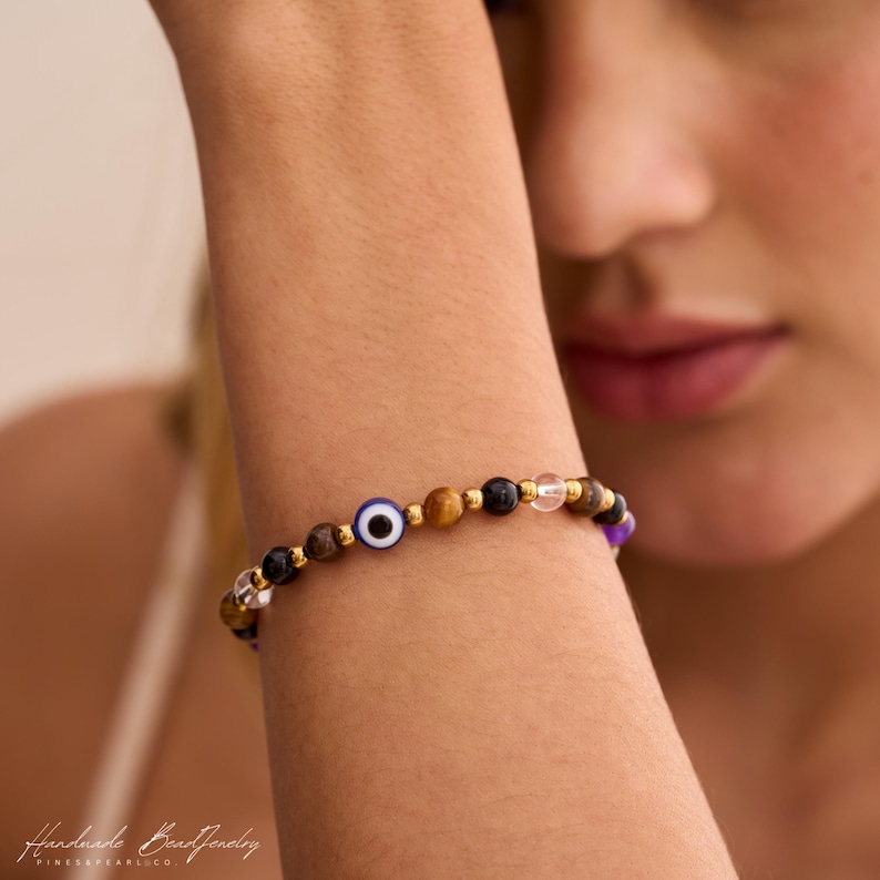 May include: A beaded bracelet featuring a central blue, white, and black evil eye charm, interspersed with polished brown tiger eye, black, clear, and purple beads, accented by small gold-toned metallic spacers throughout the circular design.