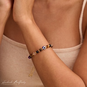 May include: A beaded bracelet featuring a central blue and white evil eye charm, interspersed with small gold-toned metal spacers and various natural stone beads in shades of brown, black, and translucent purple, with adjustable gold-toned dangling ends.