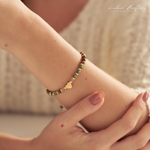 May include: A beaded bracelet featuring small, multi-colored stone beads in shades of green, brown, and pink, interspersed with tiny gold-toned metallic spacers and a central gold-toned heart-shaped charm. The bracelet is worn on a wrist.