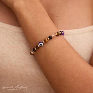 May include: A beaded bracelet featuring a mix of brown tiger eye, black, clear, and purple beads, interspersed with small gold-toned metallic spacers and a single blue, white, and black evil eye bead. The bracelet is worn on a wrist.