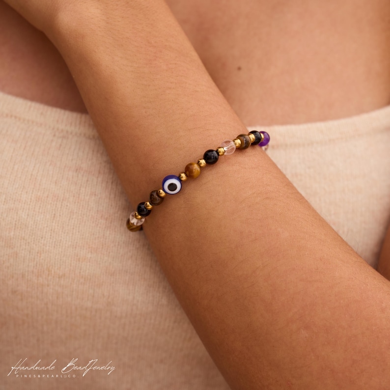 May include: A beaded bracelet featuring a mix of brown tiger eye, black, clear, and purple beads, interspersed with small gold-toned metallic spacers and a single blue, white, and black evil eye bead. The bracelet is worn on a wrist.