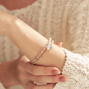 Puede incluir: Una pulsera de doble hilo que presenta un hilo de pequeñas cuentas grises translúcidas y un segundo hilo de alambre dorado trenzado con una piedra central única, iridiscente y en forma de lágrima. El texto "Handmade Bead Jewelry PINES & PEARL CO" es visible.