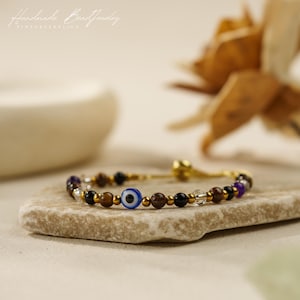 May include: A beaded bracelet featuring a central blue and white evil eye bead, interspersed with brown, black, clear, and purple spherical beads, accented by small metallic gold-toned spacer beads on a thin gold-toned chain.