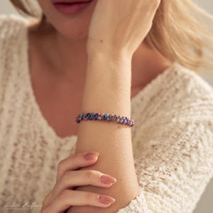 May include: A bracelet featuring a series of small, round beads in shades of purple, blue, and brown, woven together with thin, dark-colored cord. The beads have a polished, multi-colored finish that creates a vibrant, speckled appearance on the wrist.
