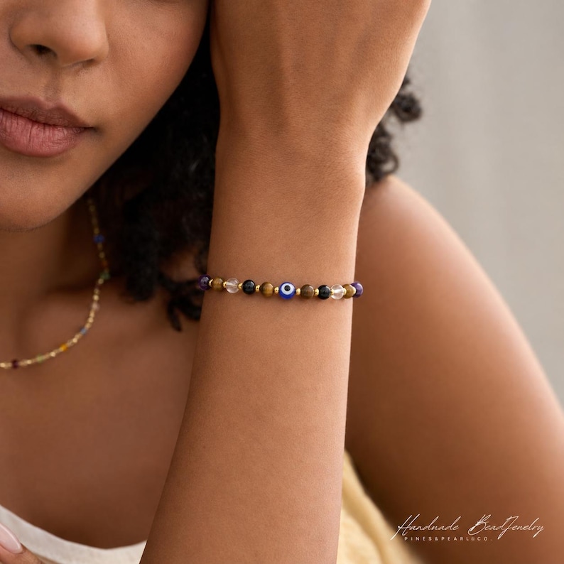 May include: A beaded bracelet featuring alternating brown tiger eye, clear quartz, and dark blue beads, accented with small gold-toned spacers and a central blue and white evil eye charm. The bracelet is worn on a wrist, showcasing a delicate design.
