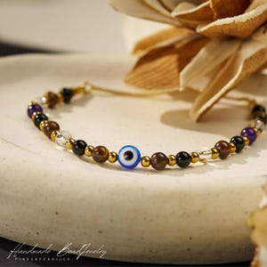 May include: A beaded bracelet featuring a central blue, white, and black evil eye bead, interspersed with brown tiger eye, black onyx, clear quartz, purple amethyst, and small metallic gold-tone spacer beads on a thin gold-tone chain.