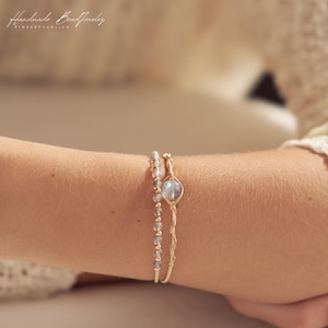 May include: Two bracelets on a wrist. One bracelet features small, round, translucent grey beads. The second bracelet is a thin gold-tone wire with a twisted design and a single, round, iridescent moonstone-like cabochon center piece.