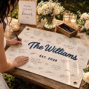 May include: A rectangular off-white fabric banner featuring the embroidered dark blue text The Williams and EST. 2026, surrounded by handwritten guest signatures and messages. The banner has metal grommets in the corners for hanging.