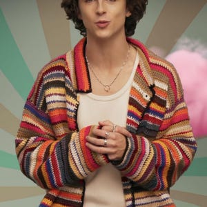 May include: A colorful, striped crochet cardigan in shades of red, orange, yellow, blue, and brown. The cardigan is worn over a white tank top and paired with a silver necklace and rings. The background features a retro-style design.