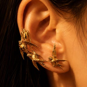 Puede incluir: Un pendiente tipo ear cuff en tono dorado que presenta un grupo de cuatro golondrinas esculpidas en vuelo. Las aves están dispuestas a lo largo del borde exterior de la oreja, con alas y cuerpos detallados que siguen la curva natural.