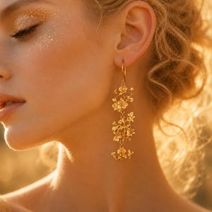 May include: A long gold dangle earring featuring a vertical arrangement of three detailed rose flower motifs connected by thin metal links. The earring has a hook-style closure and a polished metallic finish, hanging down from the earlobe.