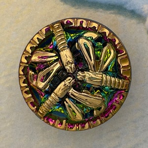 May include: A round, iridescent button with a gold-colored design of overlapping, stylized leaves. The button has a pink rim and a black center.