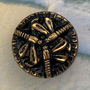 May include: A black and gold round button with a raised dragonfly design. The dragonfly has gold wings and a black body.