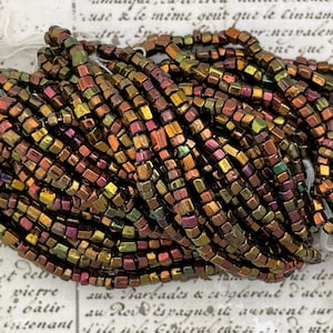 May include: A close-up of a pile of iridescent, square-shaped glass beads. The beads are a mix of brown, gold, and green colors.
