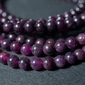 May include: Close-up of several strands of purple gemstone beads. The beads are round and have a speckled appearance, with variations in color from deep purple to lighter shades. The beads are strung together, creating a necklace or bracelet.